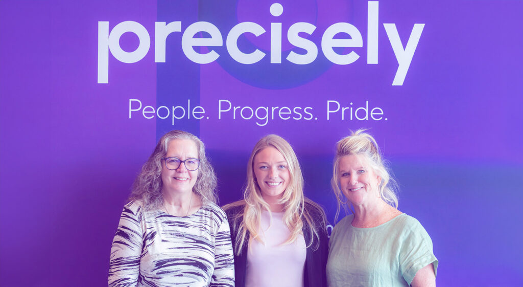 Three women employees of Precisely stand in front of the office sign.